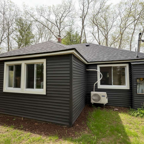 A small, dark-colored house with white-trimmed windows, surrounded by trees, with an air conditioning unit on the side.