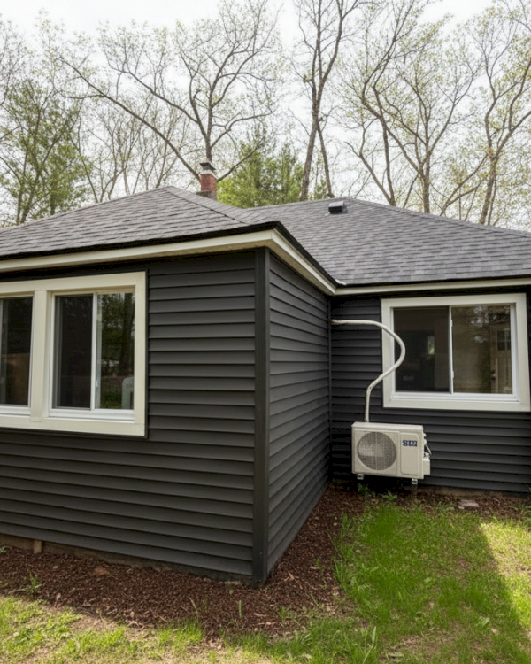A small, dark-colored house with white-trimmed windows, surrounded by trees, with an air conditioning unit on the side.