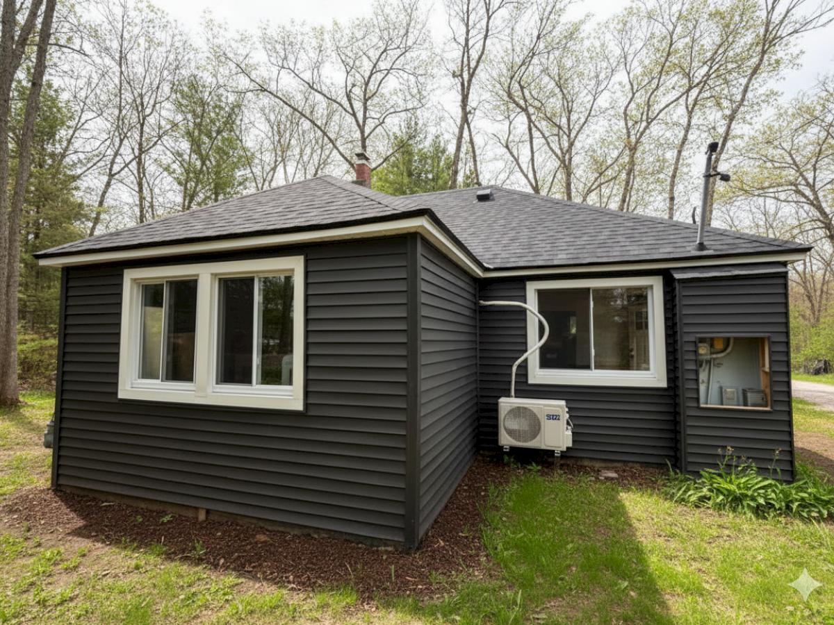 A small, dark-colored house with white-trimmed windows set in a green, wooded area, featuring an exterior air conditioning unit.